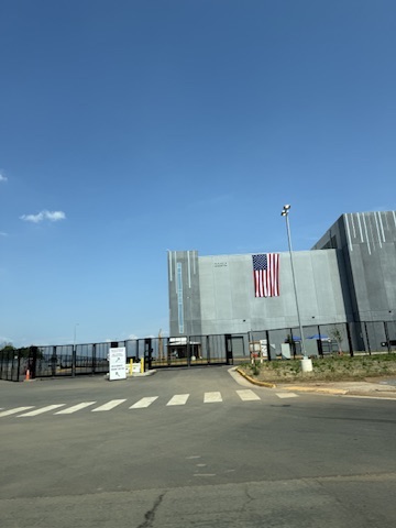 A modern data center behind a security fence on a sunny day in Loudoun County, with an American flag on the facade.