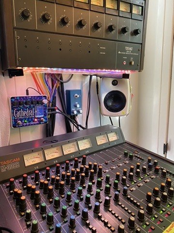 A Tascam M-308B mixing console seen up close, rows of channel faders and colored knobs, with a TEAC rack unit and a small monitor speaker behind it.