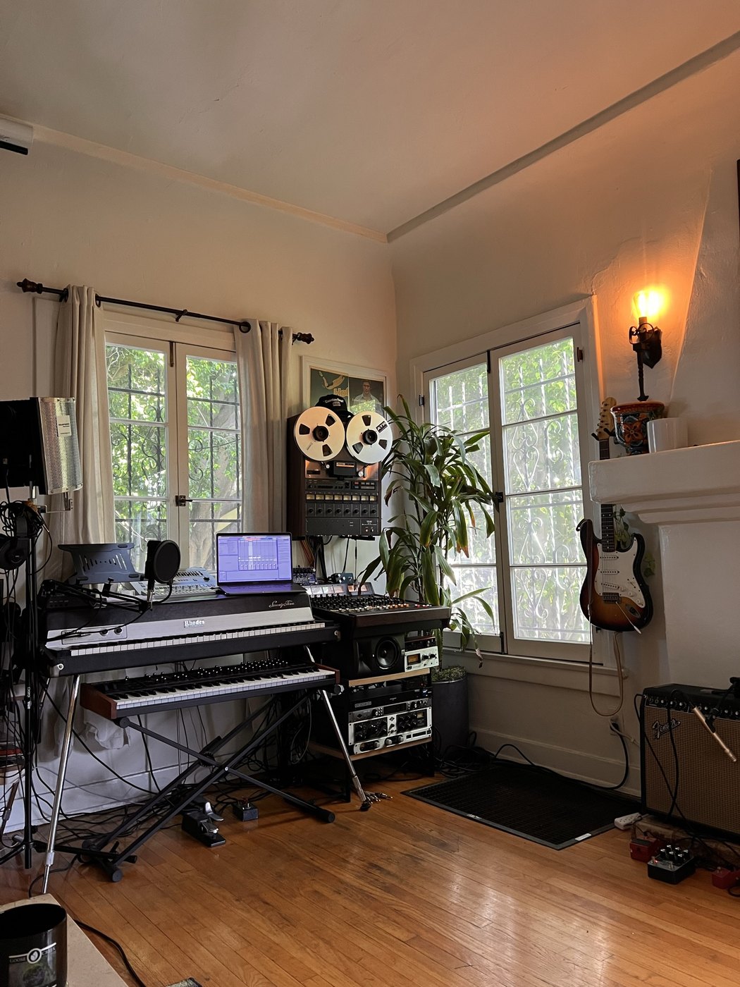A home studio with a Tascam 80-8 reel-to-reel tape machine on a stand by the window, two stacked synthesizers, a plant, and a guitar against the wall.